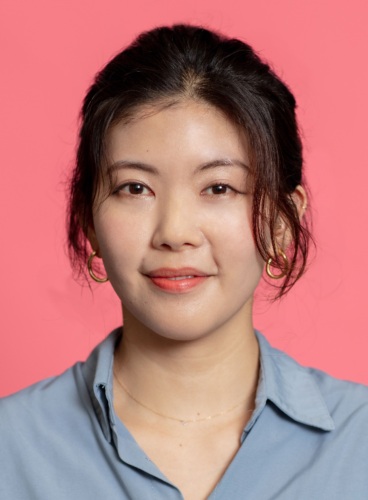 Portrait of a person with dark hair pulled back, wearing a blue shirt and gold hoop earrings, against a pink background.